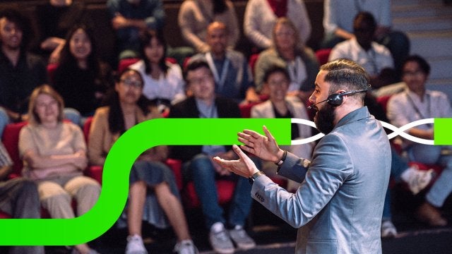 Speaker presentando frente a una audiencia en un evento corporativo, con asistentes atentos en un auditorio durante una experiencia en vivo.